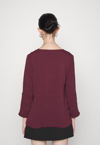 Haut à manches longues bordeaux en tissu texturé, doté d'un décolleté large et de détails subtils au niveau des poignets, porté avec une jupe noire.