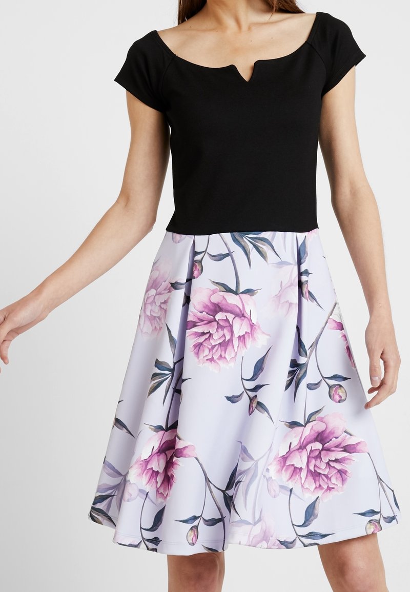 Haut noir ajusté à manches courtes avec un décolleté en cœur, associé à une jupe blanche longueur genou ornée de grandes impressions florales roses.