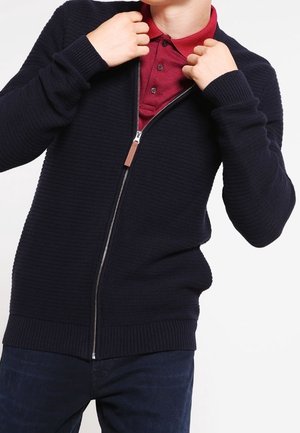 Cardigan en tricot bleu marine avec un motif texturé, fermeture éclair sur le devant et poignets côtelés. Porté par-dessus un polo bordeaux.