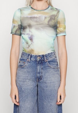 Person iført en kortærmet, tætsiddende top med et abstrakt månemotiv med skyer, kombineret med højtsiddende, blå denimjeans.