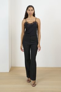 Camisole nera con dettagli in pizzo sul colletto abbinata a pantaloni neri su misura. La modella è in piedi su un pavimento di legno chiaro contro uno sfondo bianco.