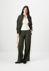 Veste en velours côtelé vert olive sur un top à côtes blanc, assortie à un pantalon olive à jambe large. Le mannequin tient un petit sac blanc.