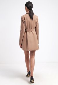 Kvinde med mørk hestehale iført en beige bæltestøvjakke og sorte højhælede sko, der går væk fra en ensfarvet hvid baggrund.