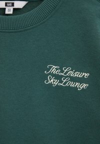 Sweat-shirt vert foncé avec texte blanc brodé "The Leisure Sky Lounge" sur la poitrine et une petite étiquette de taille à l'intérieur du col.