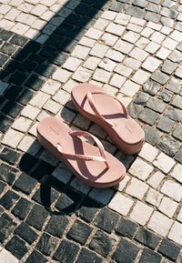 Een paar roze platformslippers die rusten op een zwart-wit geruite stenen bestrating met patronen van zonlicht en schaduw.