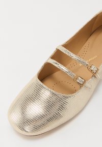 Goldene metallische Mary Jane-Schuhe mit einer strukturierten Oberfläche, die über zwei dekorative Riemen mit Schnallen verfügen und einen gepolsterten Innenraum bieten.