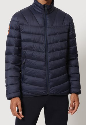 Mężczyzna w granatowej pikowanej kurtce puchowej z wysokim kołnierzem i kieszeniami na zamek, stojący na jednolitym tle.