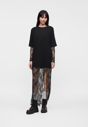 Ung kvinde stående iført en sort oversized kortærmet top lagdelt over en gennemsigtig sort blonder lang kjole med støvler og hvide sokker.