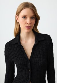 Zwarte gestructureerde shirt met een kraag, lange mouwen en een knoopsluiting. De stof heeft een gekreukte afwerking voor extra detail.