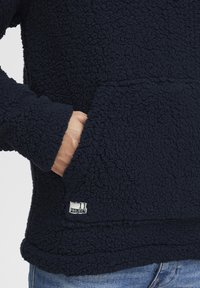 Marineblå fleece-pullover med en teksturert, fluffy overflate, kengurulomme og liten logoetikett ved kanten. Avslappet passform med lange ermer.