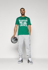 Zielona bawełniana koszulka z nadrukiem "BOSTON CELTICS", szare dresowe spodnie z bocznymi kieszeniami, trzymający czarno-szary kosz, w czarnych sneakersach.
