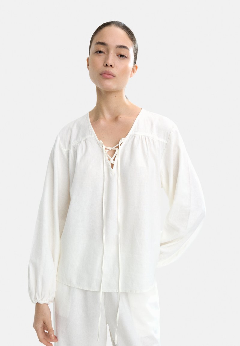 Biała bluzka z długim rękawem uszyta z lekkiego materiału, z dekoltem w kształcie litery V z ozdobnym sznurkiem, marszczonymi ramionami i elastycznymi mankietami.