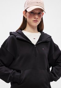 Μαύρο φούτερ με φερμουάρ και μπροστινή τσέπη, με ριμπ μανσέτες. Φοράει πάνω από ένα ανοιχτόχρωμο μπλουζάκι που διαθέτει ένα μικρό λογότυπο. Συνδυασμένο με ένα ανοιχτό ροζ καπέλο.