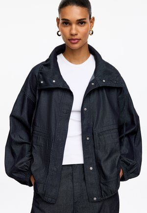 Femme portant une veste oversize sombre avec des boutons-pression sur une chemise blanche, les mains dans les poches et des boucles d'oreilles créoles dorées.