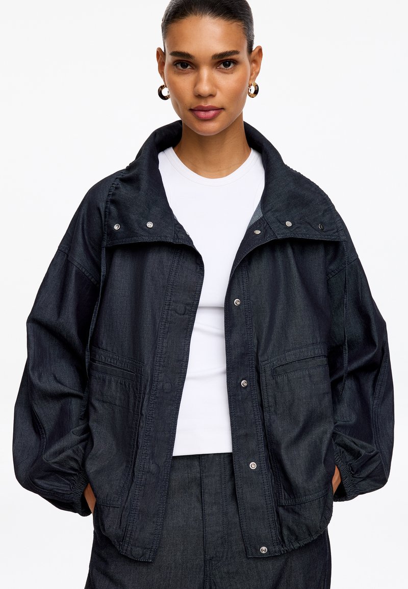 Femme portant une veste oversize sombre avec des boutons-pression sur une chemise blanche, les mains dans les poches et des boucles d'oreilles créoles dorées.