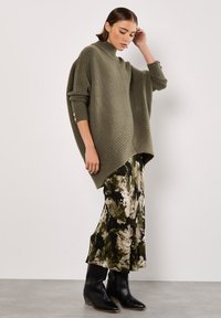 Dzianinowy, oliwkowozielony, oversize'owy sweter z wysokim kołnierzem i prążkowaną teksturą, zestawiony z czarną i oliwkową spódnicą maxi w kwiaty oraz czarnymi botkami za kostkę.