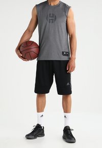 Chemise grise sans manches avec un logo, short noir, chaussures de sport noires et chaussettes blanches. Tenant un ballon de basket, debout devant un fond uni.