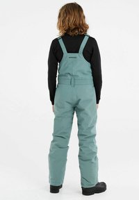 Salopette impermeable vert menthe avec bretelles ajustables, taille élastique et tissu durable. Haut en tissu noir en dessous et bottes noires visibles.