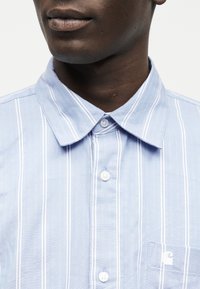 Κλείσιμο εικόνας ενός ατόμου που φοράει πουκάμισο με κάθετες ρίγες σε γαλάζιο και λευκό χρώμα, με κουμπιά, γιακά και μικρό λογότυπο στην τσέπη.