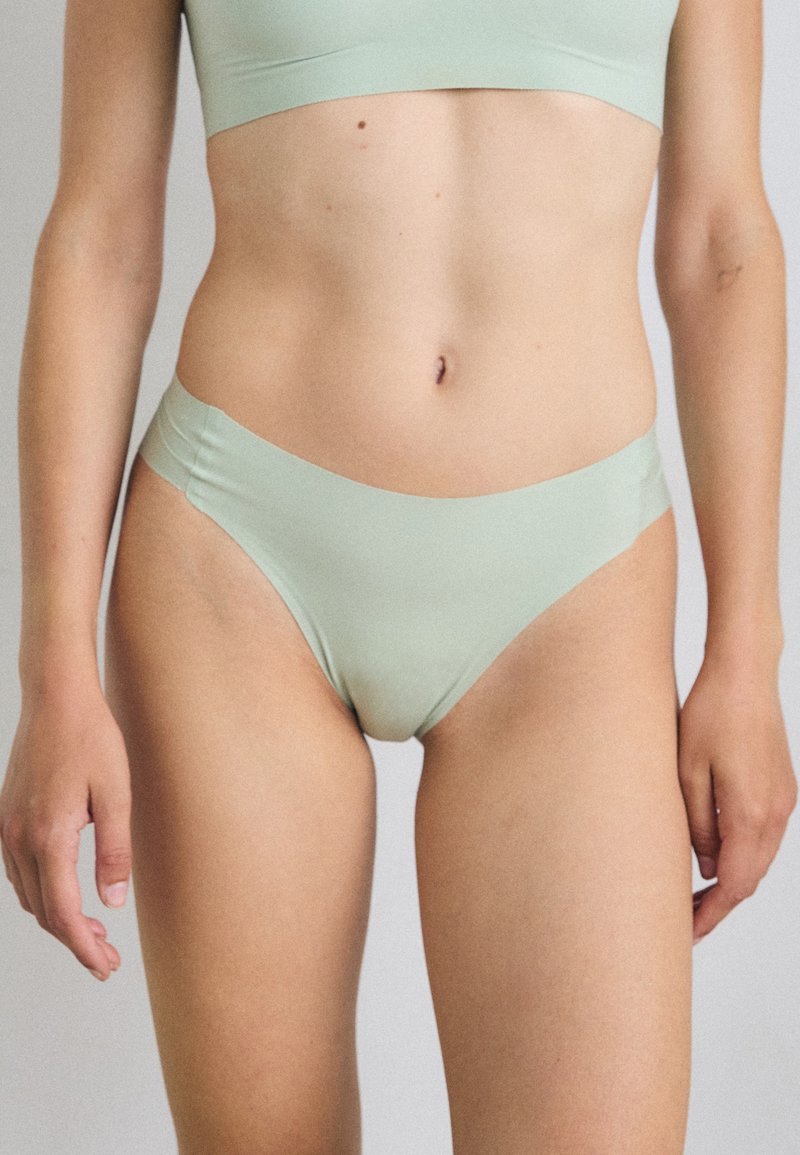 Modello che indossa un bralette verde chiaro e delle mutandine in stile bikini, in piedi contro uno sfondo neutro, con le braccia rilassate lungo i fianchi.