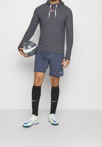 Nike Langarm-Hoodie in Dunkelgrau mit kontrastierendem orangen Logo. Kombiniert mit marineblauen Shorts, schwarzen Knielangen Socken und weißen Fußballschuhen mit blauen Akzenten.