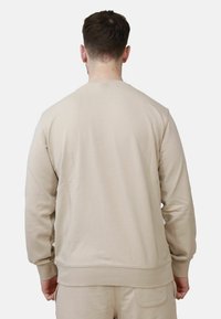 Man in een effen beige sweatshirt met lange mouwen en bijpassende beige broek, gezien vanaf de achterkant tegen een witte achtergrond.