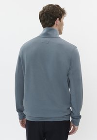 Szürke, hosszú ujjú pulóver, magas gallérral, puha anyagból készült. Minimalista varrásokkal és laza szabással rendelkezik.