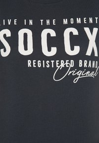 Camiseta negra de algodón con gran texto impreso en blanco: "VIVE EL MOMENTO", "SOCCX" y "MARCA REGISTRADA Original."
