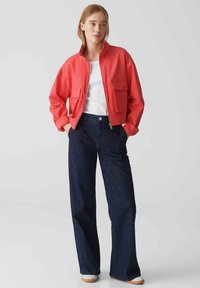 Rote Bomberjacke mit Reißverschluss und zwei vorderen Taschen, getragen über einem weißen Hemd und blauen weit geschnittenen Jeans, vor einem neutralen Hintergrund präsentiert.