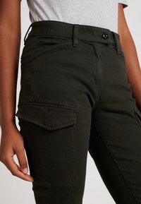 Cargo pants made of dark green fabric with a fitted design, featuring side pockets, a button closure, and visible stitching details.