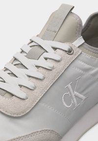 Κοντινό πλάνο ανοιχτόγριου και μπεζ αθλητικού παπουτσιού με λευκά κορδόνια και κέντημα με το λογότυπο «ck» στην πλευρά κοντά στον αστράγαλο.