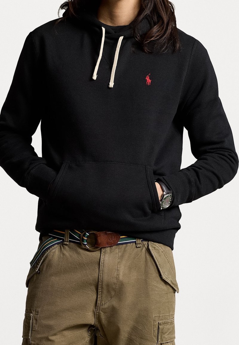 Svart huvtröja med framficka, prydd med en röd logotyp, beige dragsko och ribbade muddar, ihop med khaki cargobyxor och ett brunt bälte.