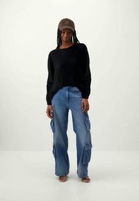 Femeie îmbrăcată cu o pălărie maro, pulover negru din tricot, blugi cargo cu picioare largi de culoare albastră și sandale cu degete goale, stând pe un fundal simplu.