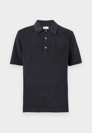 Homme aux cheveux blonds courts portant un polo noir par-dessus un t-shirt rayé, un jean bleu ample et des chaussures noires, debout avec une main dans la poche.