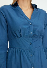 Donna che indossa un vestito blu a maniche lunghe con bottoni, caratterizzato da una cintura ampia e leggere arricciature sotto la vita.