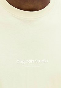 T-shirt beige clair avec un col rond. Dispose d'une texture douce et d'un imprimé blanc "Originals Studio" sur le devant.
