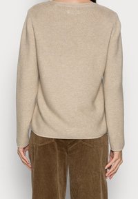 Person, der einen beigefarbenen Strickpullover und eine braune Cordhose trägt, von hinten gesehen mit entspannten Armen an den Seiten.