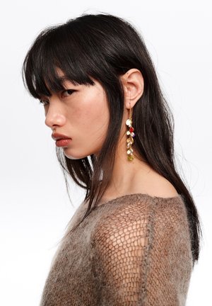 Młoda kobieta o długich ciemnych włosach, ubrana w przezroczysty brązowy sweter z dzianiny i długi złoty kolczyk z kolorowymi koralikami, patrząca na bok.
