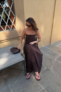 Braunes, plissiertes Maxikleid mit dünnen Trägern, kombiniert mit einer strukturierten Handtasche. Das Model sitzt auf einem Stuhl und trägt Flip-Flops auf einem steinernen Boden.