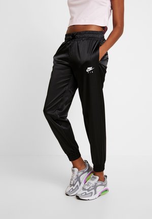 Czarne spodnie dresowe Nike Air z elastycznymi mankietami i pasem, noszone z białymi, szarymi i zielonymi sneakersami sportowymi na stojącej osobie.