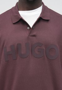 Close-up af en person iført en bordeaux polo med store, hævede bogstaver, der staver "HUGO" tværs over brystet.