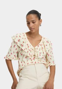 Lichtgele bloemenblouse met gerimpelde mouwen, V-hals en een strik aan de voorkant, versierd met roze rozenpatronen. Zachte stoftextuur.