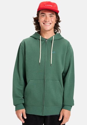 Green zip-up hoodie made of soft fabric, featuring a kangaroo pocket, drawstring hood, and a small logo accent. Worn with a red cap.