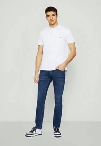 Biela polo tričko s krátkymi rukávmi v kombinácii s tmavomodrými džínsy. Model má na sebe čierno-biele tenisky a stojí s jednou rukou v vrecku.