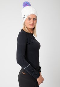 White knitted beanie with a colorful pom-pom on top, worn with a black long-sleeve top featuring textured fabric and a visible logo.