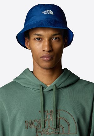 Niebieska czapka z daszkiem o teksturowanym materiale z białym logo "The North Face". Noszona z zieloną bluzą z kapturem, prezentując swobodny wygląd.