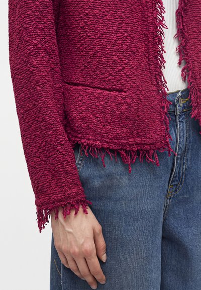 Personne portant une veste rouge en bouclé texturée avec bords à franges, un jean bleu et un t-shirt blanc côtelé visible en dessous. Main relâchée le long du corps.