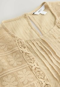 Beige blouse, millel plekitud disain, keerukad pitsaktsendid ja võtmesulgur taga. Valmistatud kerge tekstureeritud kangast.
