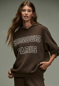 Felpa con cappuccio marrone che presenta la scritta "BISOU PARIS" in caratteri bianchi, con vestibilità ampia e polsini a coste, realizzata in un morbido tessuto testurizzato.