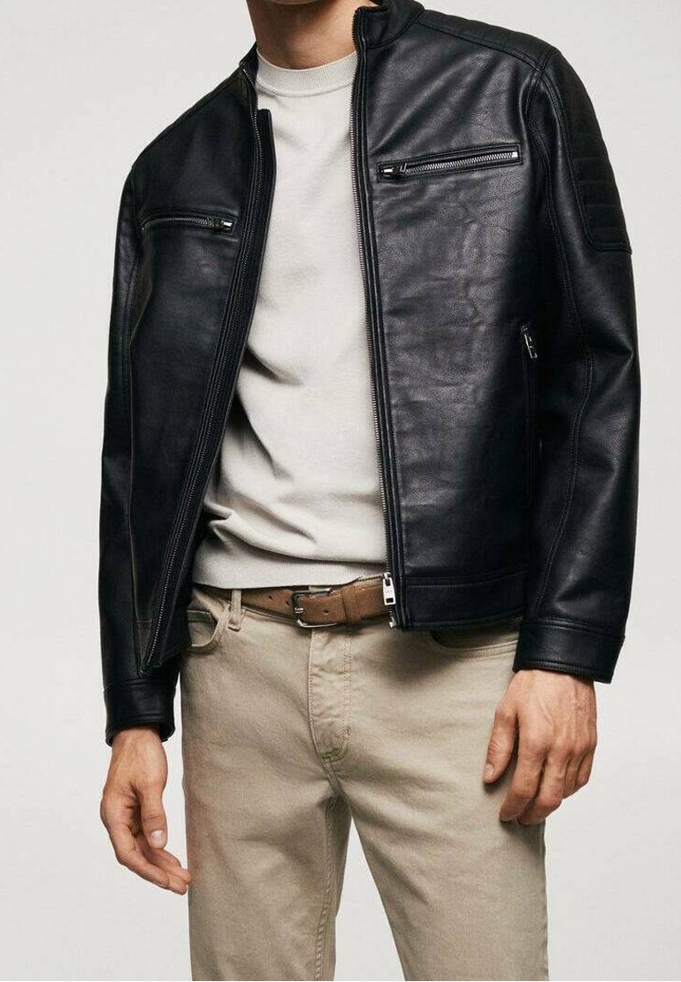 Homme portant une veste en cuir noire, un pantalon beige, une ceinture marron et un t-shirt beige clair à col rond, debout les mains détendues le long du corps.